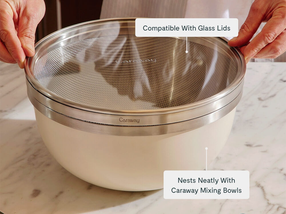 Hands fitting a glass lid over a stainless steel strainer nested in a Cream mixing bowl.