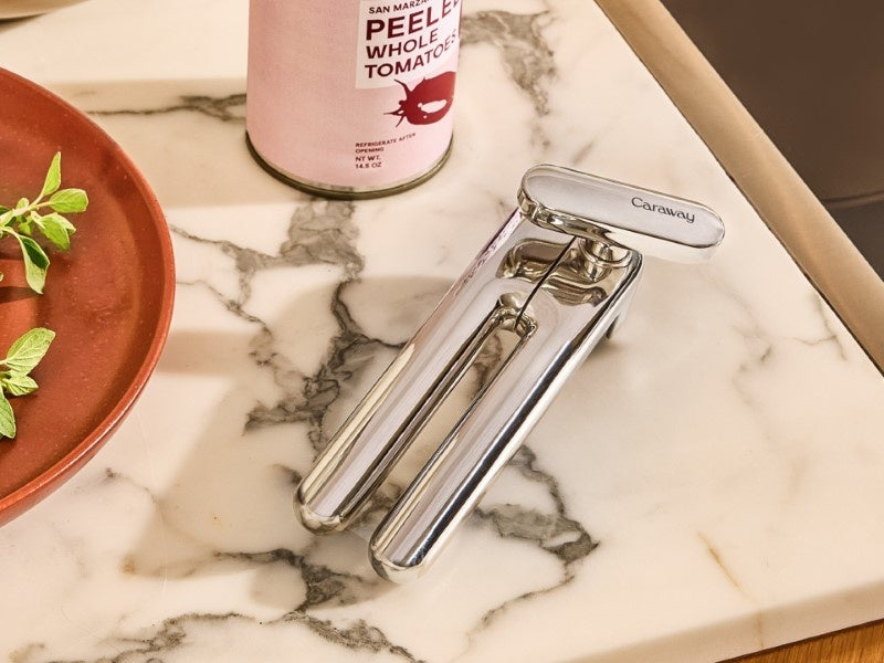 Caraway stainless steel can opener beside a can of tomatoes on a kitchen counter.