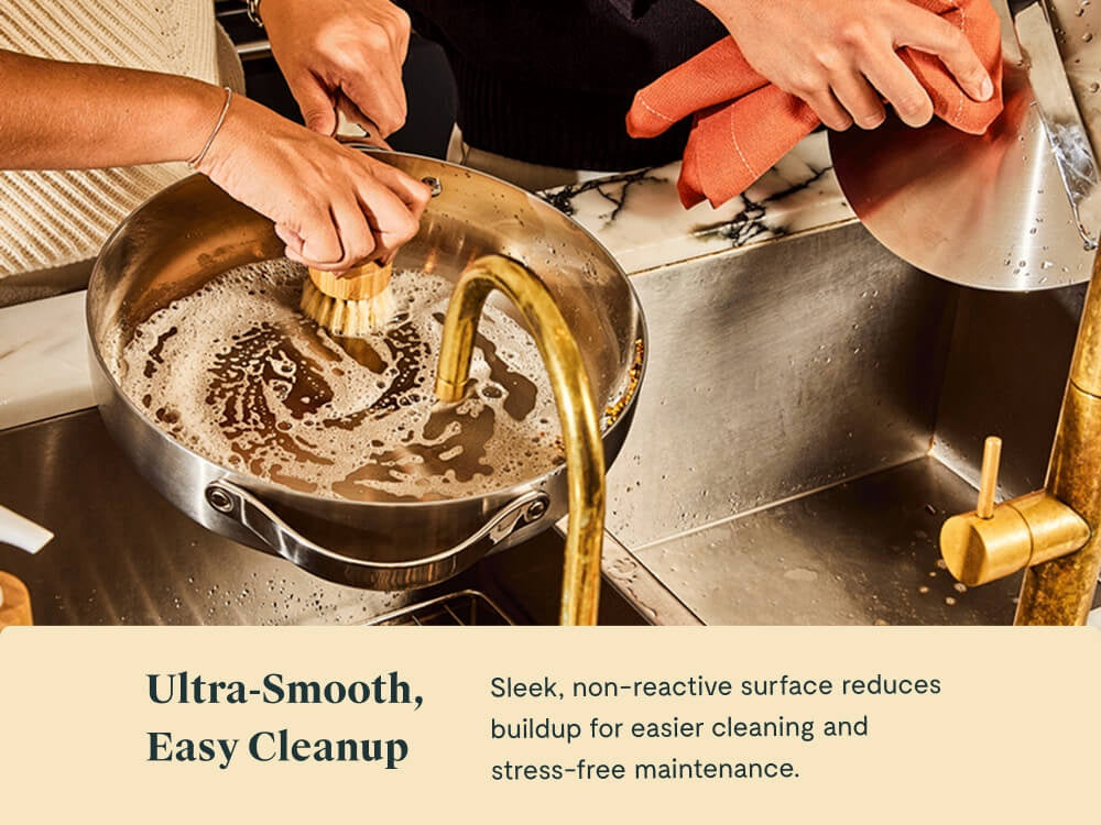 Hands washing stainless steel cookware with a brush and soapy water in a sink.