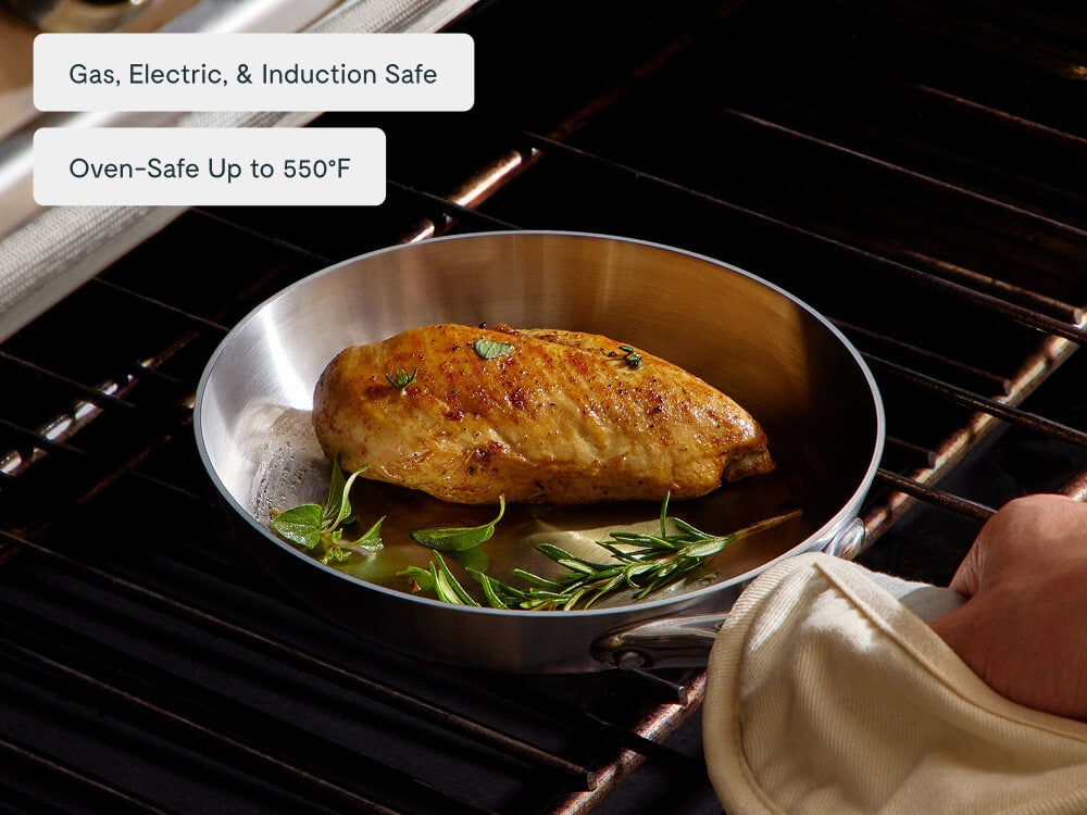 Hand with a towel removing a stainless steel fry pan of roasted chicken from an oven.