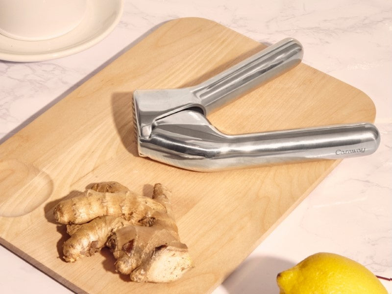Caraway stainless steel garlic press beside ginger on a wooden cutting board.