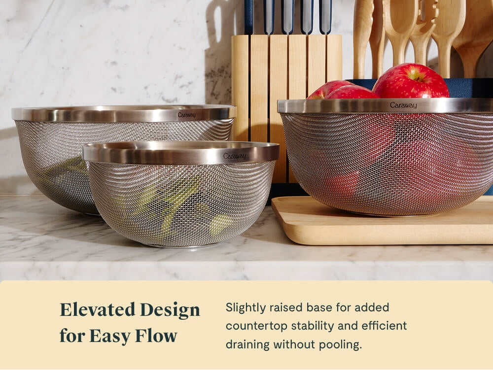 Caraway stainless steel strainers in different sizes holding produce on a kitchen counter.