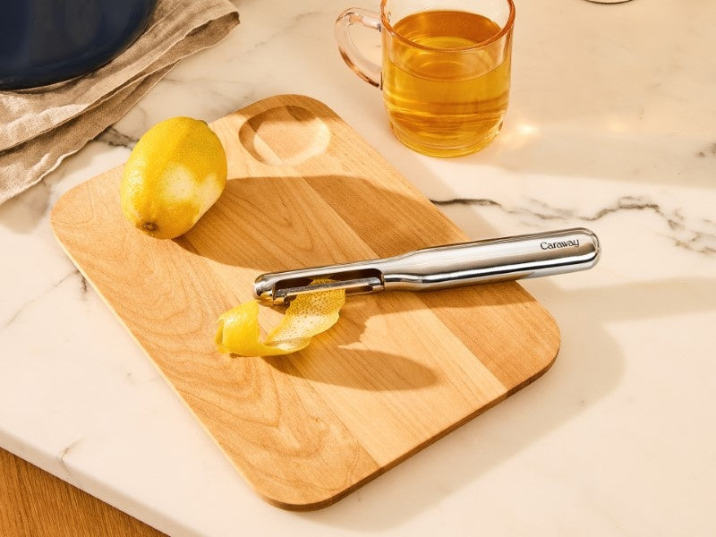 Caraway stainless steel vegetable peeler beside lemon peel on a wooden cutting board.