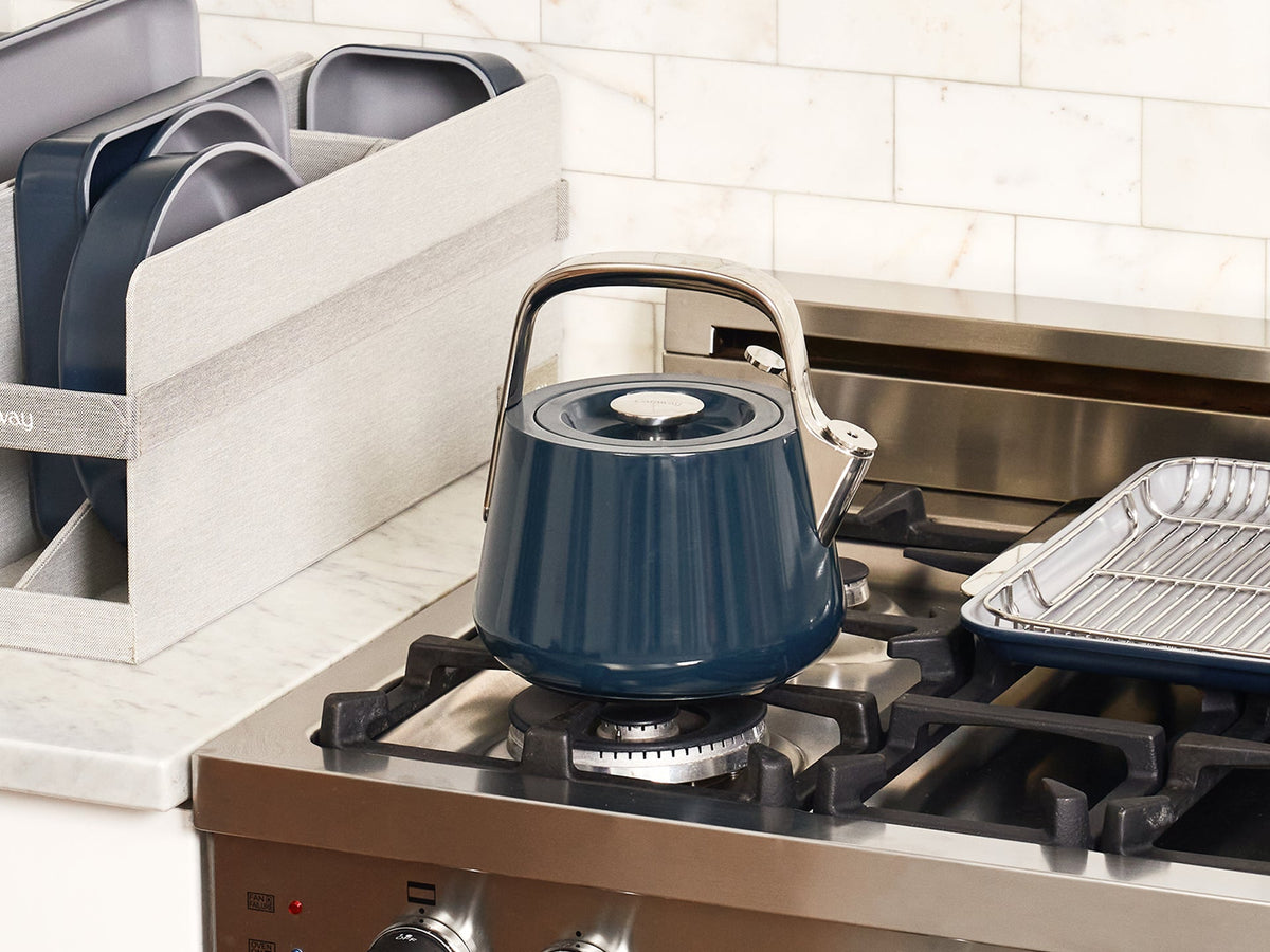 Caraway Navy whistling tea kettle beside a stove on a kitchen counter.