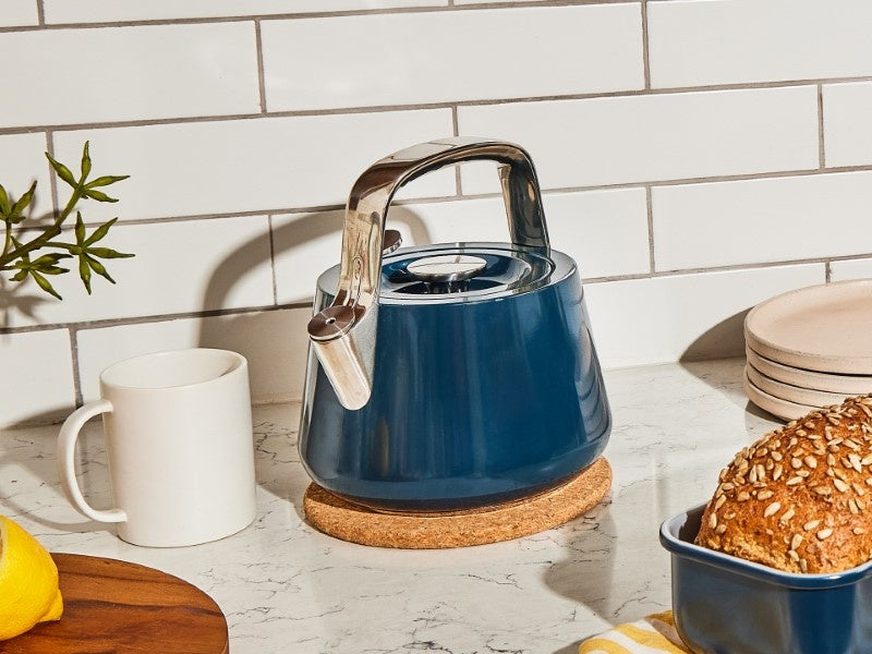 Caraway Navy whistling tea kettle beside a stove on a kitchen counter.